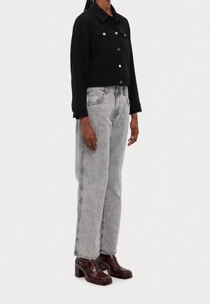 Person iført en sort cropped jakke, lysegrå højtaljede jeans og mørkebrune ankelstøvler med hæl, stående mod en ensfarvet baggrund.