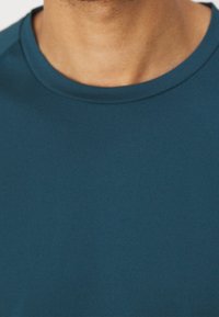 Tyrkysové tričko s kulatým výstřihem vyrobené z hladké látky. Má jednobarevný design bez vzorů, což zdůrazňuje jeho jednoduchost.