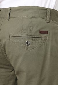 Gros plan sur la poche arrière d'un pantalon vert olive avec fermeture à bouton et petite étiquette en cuir avec la marque au-dessus de la poche.