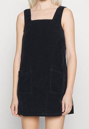 Femme portant une robe salopette en velours côtelé noir sans manches avec deux poches devant, sur un fond clair uni.