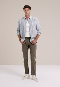 Svijetložuta i bijela prugasta košulja s dugim rukavima, bijela majica bez rukava, maslinasto zelene hlače i bijele tenisice, stoji naslonjena na neutralnu pozadinu.