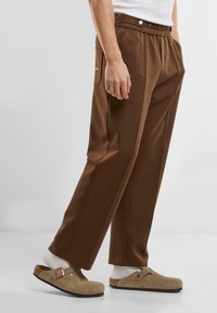 Brown relaxed-fit trousers with an elastic waistband, side pockets, and a smooth texture, paired with beige clogs and white socks.