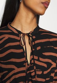 Gros plan sur une personne portant un haut noir à rayures brunes avec un décolleté en trou de serrure noué et une boucle d'oreille argentée en forme d'anneau à l'oreille gauche.