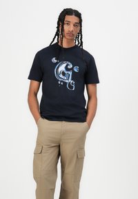 Marineblauwe katoenen T-shirt met een grote afbeelding van bellen en een gestileerde letter "G" in verschillende tinten blauw, gecombineerd met khaki cargo shorts.