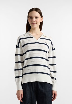 Kvinde iført hvid sweater med marineblå vandrette striber og mørke bukser, kigger let til siden med et neutralt udtryk.