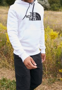 Hombre vestido con un hoodie blanco de The North Face y pantalones negros, de pie al aire libre entre plantas verdes y amarillas.
