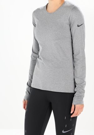Osoba ubrana w dopasowaną szarą koszulkę z długim rękawem marki Nike i czarne legginsy Nike z subtelnymi odblaskowymi paskami stoi na białym tle.