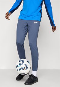 Nike treningsbukser i grått med en tettsittende design. Kombinert med en blå langermet skjorte og svarte fotballsko. Hvit fotball ved føttene.