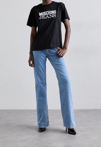 Osoba stojí v černém tričku s nápisem "MOSCHINO JEANS" vytištěným bílou barvou, má na sobě světle modré zvonové džíny a černé vysoké podpatky.