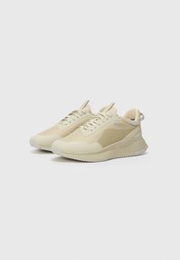 Beige athletic shoes with a textured mesh upper, smooth overlays, and a ridged rubber sole, featuring minimal branding on the side and tongue.
