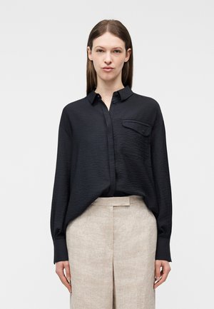 Ženska v široki črni srajci z gumbi in dolgimi rokavi ter svetlo bež hlačah z visokim pasom, stoji ob enobarvni svetli podlagi.