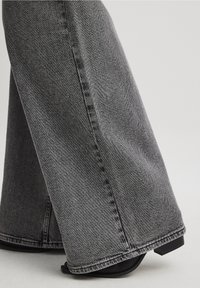 Gros plan sur un jean gris à jambes larges couvrant des chaussures à talons noires sur un fond clair.