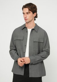 Mladý muž s vlnitými hnedými vlasmi, oblečený v sivej košeli na gombíky cez biele tričko, stojaci pred jednoduchým svetlým pozadím.