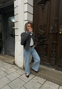 Chaqueta gris ajustada, suéter blanco texturizado, jeans de pierna ancha de color azul claro y zapatos negros. Sosteniendo una taza de café, de pie junto a una puerta de madera.