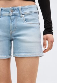 Ανοιχτό μπλε τζιν σορτς φορεμένα από ένα άτομο με μαύρο μπλουζάκι μακριάς μανίκας, που δείχνει τις μπροστινές τσέπες και το κούμπωμα με κουμπιά.