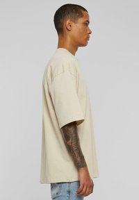 Beige oversized tričko s krátkým rukávem, kulatým výstřihem a měkkou texturou. Model má tetování na předloktí.