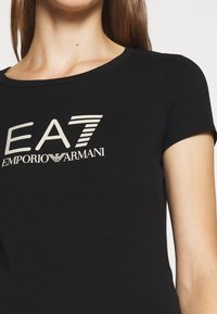 T-shirt en coton noir avec un col ronde, des manches courtes, présentant un grand graphisme en blanc et métallique incluant "EA7" et "Emporio Armani."