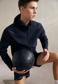 Felpa sportiva nera con zip, dettagli grigi e pantaloni abbinati. L'individuo tiene una palla medica nera texturizzata, seduto su una superficie chiara.