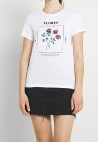 Bílá bavlněná tričko s květinovým grafickým potiskem v červené a zelené barvě, s textem "FLORES de la vida" a obdélníkovým rámem.