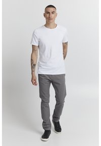 Camiseta blanca de algodón, pantalones ajustados grises y zapatillas negras. El modelo está de pie con los brazos relajados, contra un fondo claro y liso.