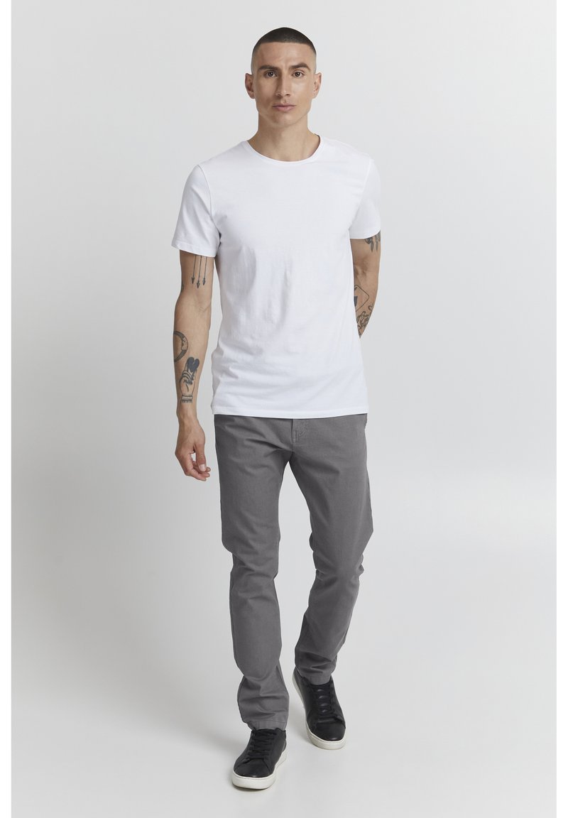 Camiseta blanca de algodón, pantalones ajustados grises y zapatillas negras. El modelo está de pie con los brazos relajados, contra un fondo claro y liso.