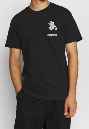 Black cotton t-shirt featuring a white cartoon character graphic and "adidas" logo on the left chest. Short sleeves and relaxed fit.