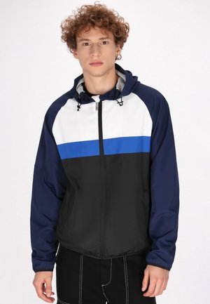 Windbreakerjack met capuchon in een kleurblokdesign in wit, blauw en zwart, met een ritssluiting en elastische manchetten. Soepel, lichtgewicht materiaal.