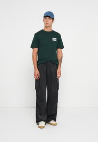 Ung mann iført en blå caps, mørkegrønn "Alpine Goods" t-skjorte, løse mørke cargo-bukser og hvite joggesko, som står mot en hvit bakgrunn.