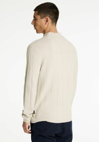 Joven en suéter crema de canalé, de pie de lado con la cabeza girada, usando pantalones oscuros frente a un fondo blanco liso.