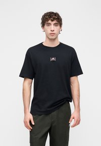 T-shirt preta de algodão com gola redonda, apresentando um pequeno emblema gráfico rosa no peito. Corte amplo e mangas curtas.