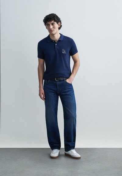 Μπλε ναυτικό πόλο μπλουζάκι με κοντά μανίκια και γιακά, διακοσμημένο με κεντημένο λογότυπο. Συνδυασμένο με μπλε τζιν παντελόνι και λευκά αθλητικά παπούτσια.
