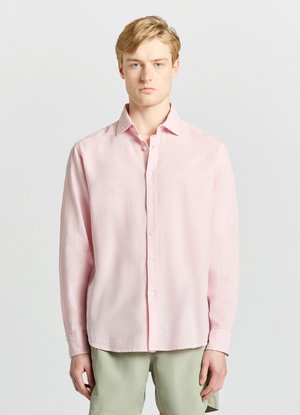 Jeune homme face à l'objectif portant une chemise rose clair à manches longues boutonnée et un short vert clair sur un fond uni.