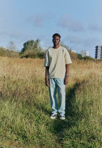 Sweat-shirt beige surdimensionné à manches courtes, jean bleu clair, et baskets noires/blanches ; debout dans un champ herbeux sous un ciel dégagé.