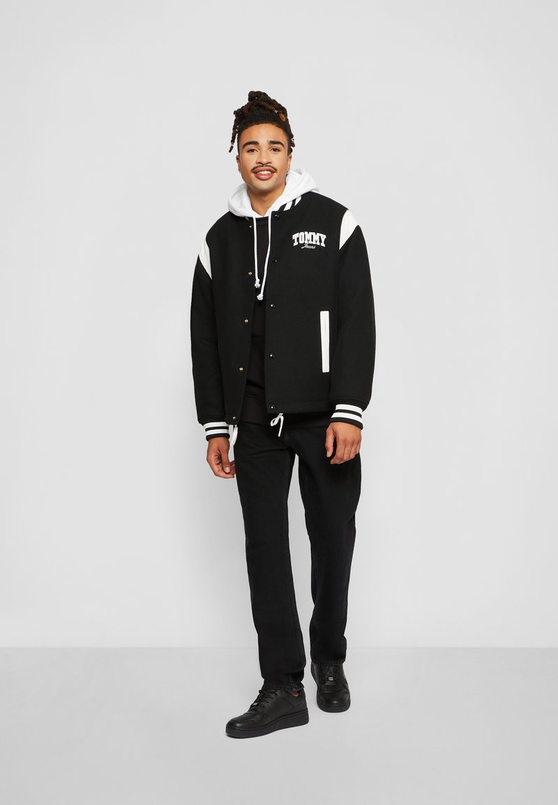Svart varsity-jakke med hvite detaljer, med en logo, ribbede mansjetter og gullknapper. Kombinert med en hettegenser, svarte bukser og svarte joggesko.