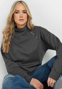 Grå sweatshirt med hög krage och dragkedjedetalj. Långa ärmar och ribbade muddar. Bärs med blå denimjeans. Mjuk textur.