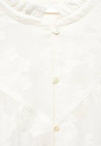 Blouse blanche texturée avec col rond à volants et trois petits boutons blancs sur le devant.