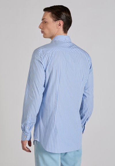 Mężczyzna w jasnoniebieskiej i białej koszuli w pionowe paski z długim rękawem, zwrócony w lewo, tyłem do kamery, ubrany w jasnoniebieskie spodnie na jednolitym tle.