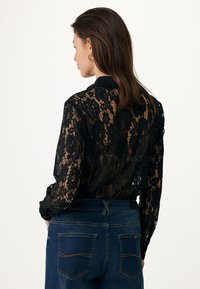 Zwarte kanten blouse met bloemenpatroon, lange mouwen en een kraag; gecombineerd met donkerblauwe denim jeans met subtiele stiksels op de zakken.