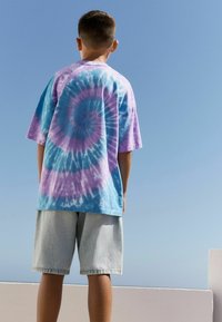 Tricou cu mânecă scurtă, tie-dye, având modele în spirală de culoare albastră, violetă și lavandă, asociat cu pantaloni şort din denim deschis.