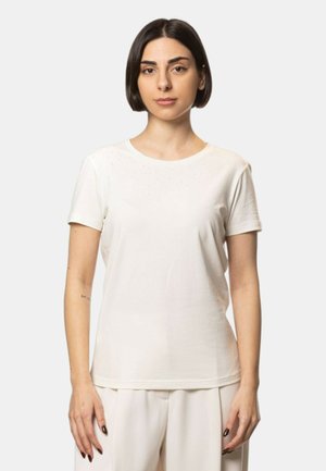 Giovane donna con capelli lisci scuri che indossa una maglietta bianca semplice a maniche corte e pantaloni chiari, in piedi davanti a uno sfondo neutro.