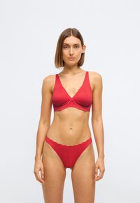 Röd underklädesuppsättning som innehåller en slät bralette med scalloped-kant och matchande stringtrosa, båda i stretchigt tyg med en subtil textur.