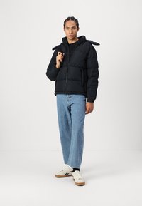 Sort polstret jakke med en frontlynlås, der har skulderstropper; båret over en sort skjorte og kombineret med lyseblå jeans og hvide sneakers.