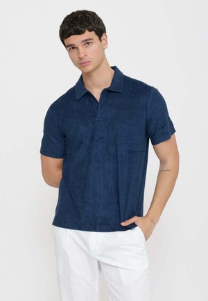 Giovane uomo con capelli corti e scuri che indossa una polo blu navy a maniche corte e pantaloni bianchi su uno sfondo chiaro e uniforme.