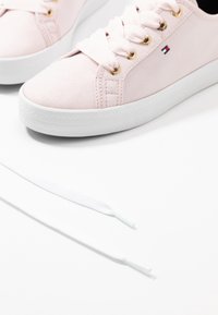 Světle růžové tenisky s hladkým povrchem, zlatými očky, bílou gumovou podrážkou a plochými bílými tkaničkami umístěnými vedle bot.