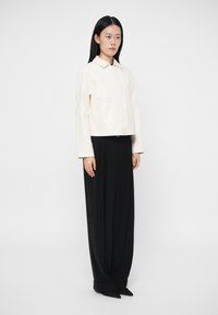 Γυναίκα που στέκεται φορώντας ένα κρεμ σακάκι με κουμπιά και μαύρους παντελόνες με φαρδιά πόδια, μπροστά σε ένα απλό λευκό φόντο.