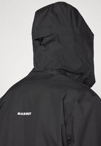 Giacca nera impermeabile con cappuccio, caratterizzata da una texture liscia e una piccola tasca. Il logo "MAMMUT" è visibile sul retro.