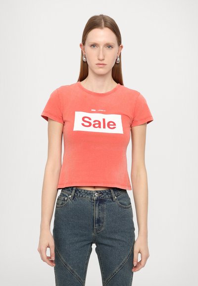 T-shirt court corail avec un graphisme blanc rectangulaire "Soldes". Matière en coton doux, manches courtes, col rond, associé à un jean taille haute.