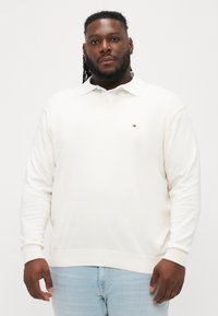 Krém színű pulóver V-nyakkal és hosszú ujjakkal. Kis logó található a mellkason, bordázott mandzsettákkal készült, puha anyagból.