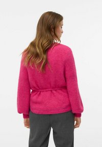 Cardigan en tricot rose avec ceinture à nouer, manches longues et finition texturée. Porté sur un pantalon gris, mettant en avant une coupe décontractée et un style casual.