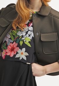 Chaqueta recortada verde oliva con cuello redondeado y dos bolsillos delanteros, superpuesta a una camisa de patrones florales que presenta vibrantes flores rojas, rosas y blancas.